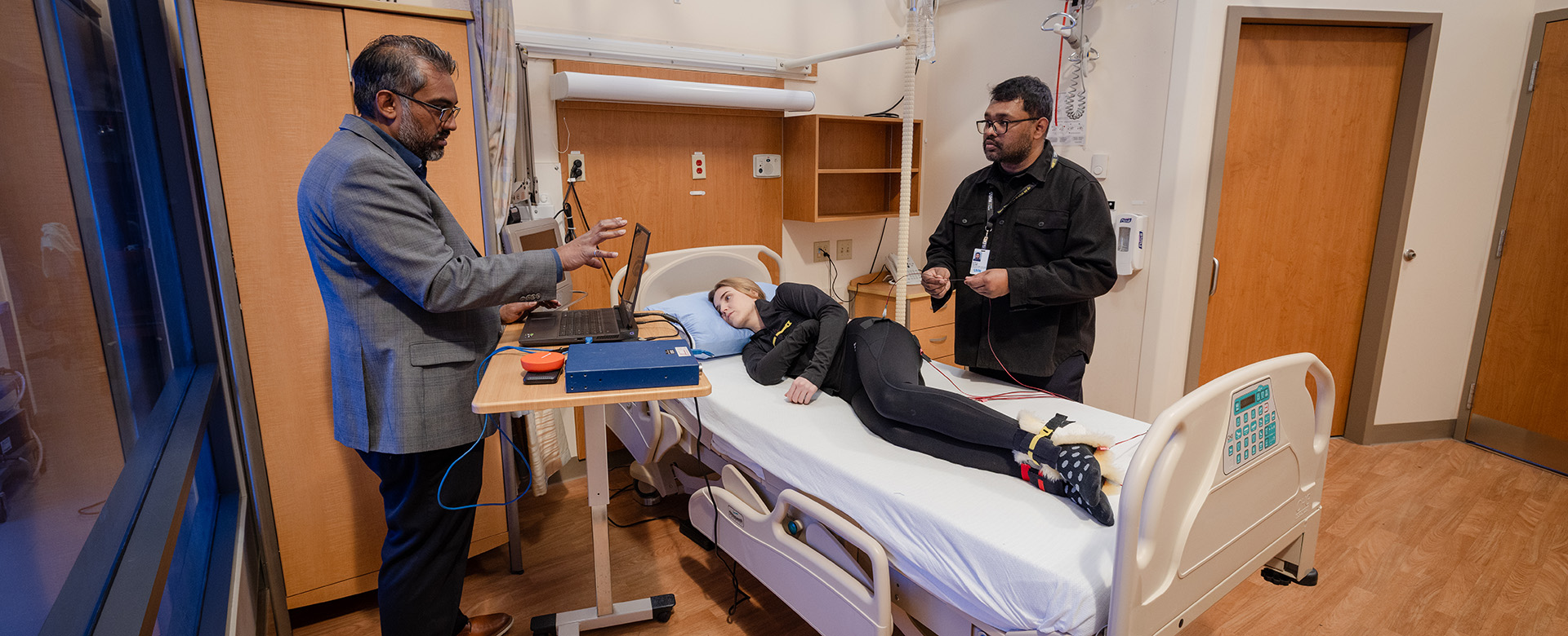 Two researchers are discussing data on a laptop, standing around a woman lying in a hospital bed with monitoring equipment.