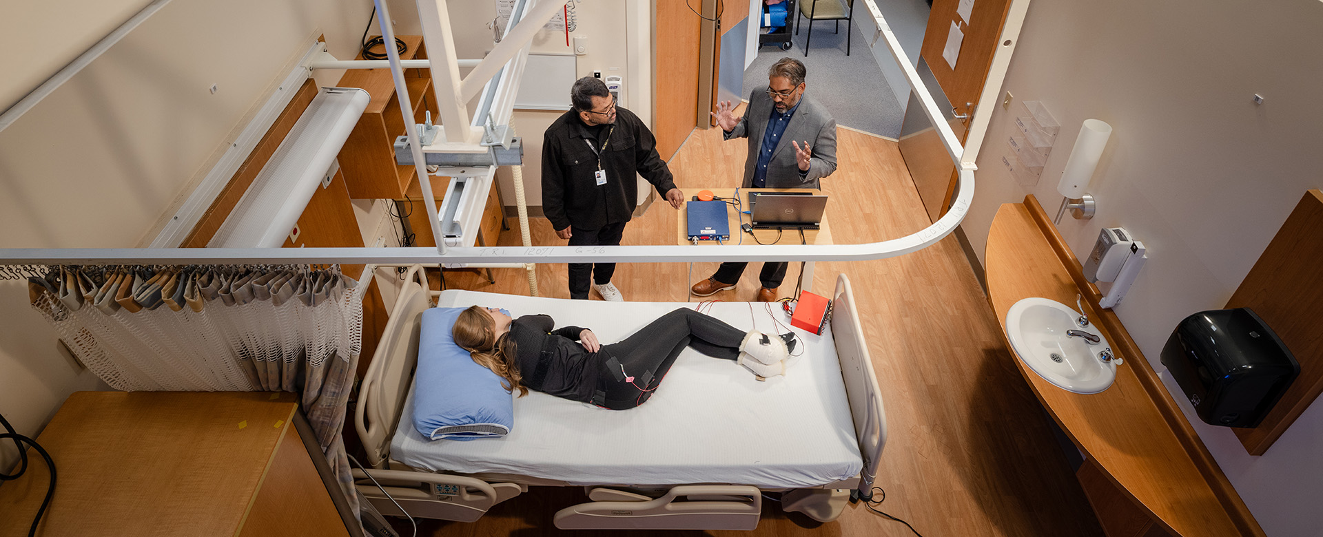 A photo taken from above, showing a hospital room with two researchers discussing data on a laptop. A patient is lying in a hospital bed with monitoring equipment around her.