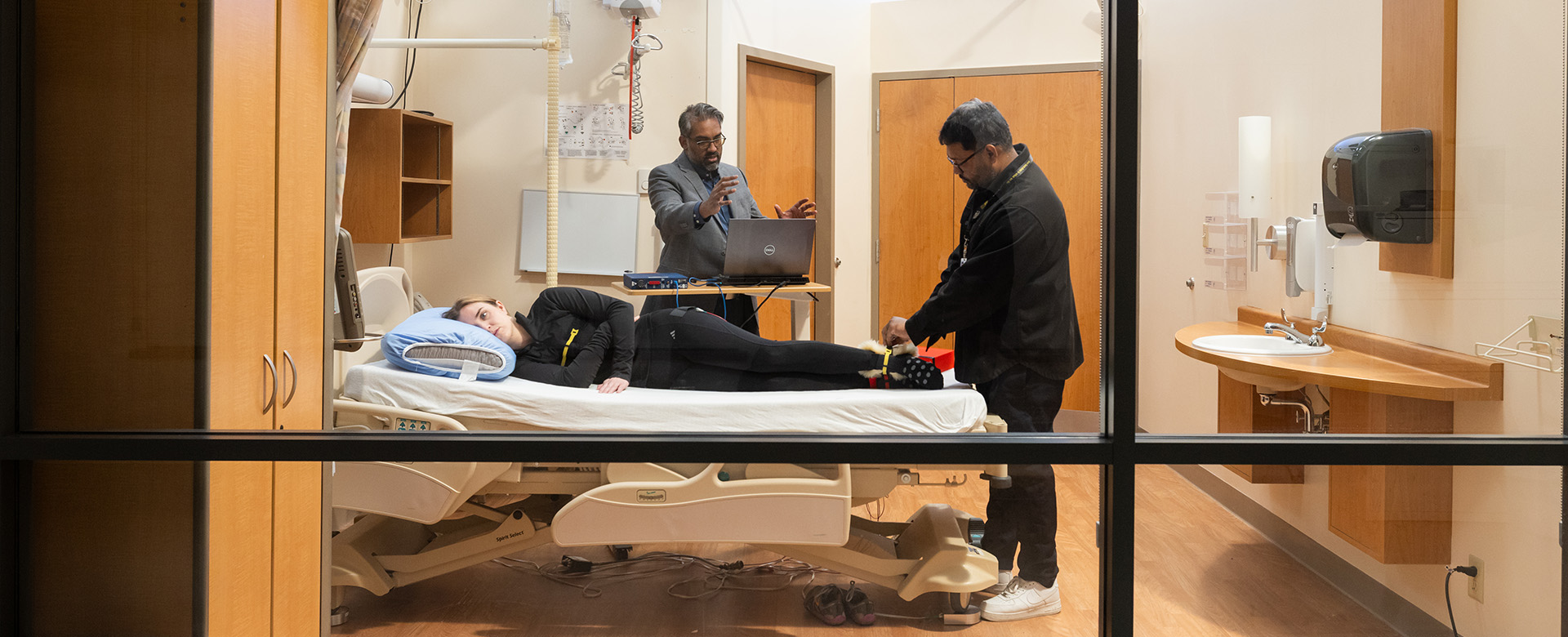 A woman is lying on a hospital bed with a researcher adjusting a sensor on her ankle. A second researcher is using a laptop in the background.